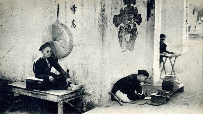 A female fortune-teller and a physiognomist practicing their trade outside a temple in Hanoi around the late 19th to early 20th century.